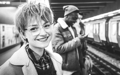 Happy young woman with glasses and a pixie haircut at a London underground station, symbolizing accessible coding education in the heart of the city.