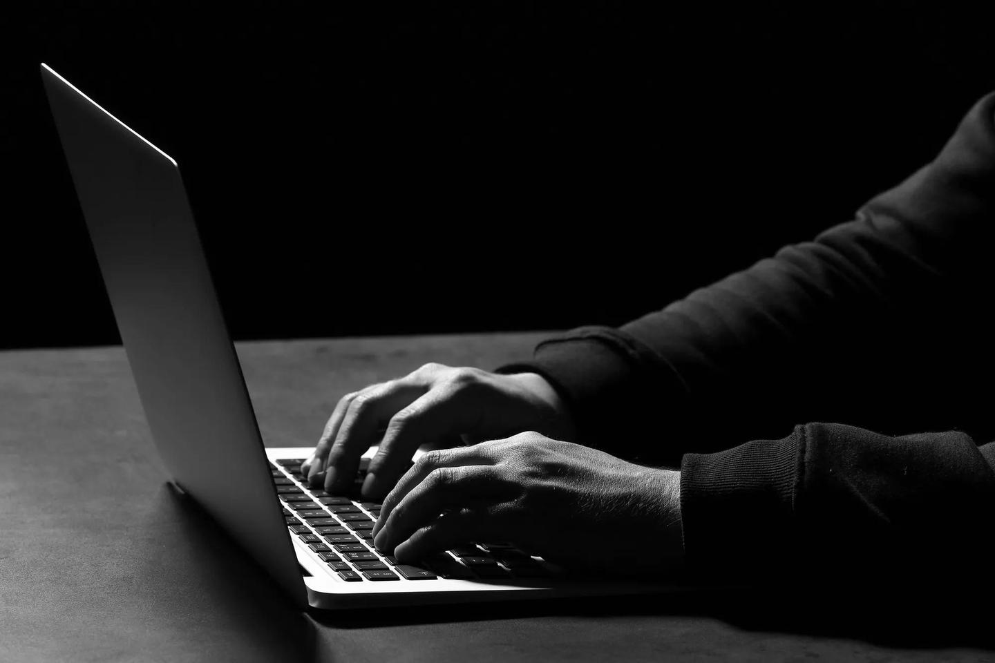 Professional hacker with laptop sitting at table on dark background, closeup