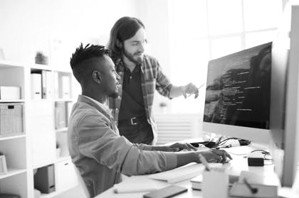 Positive skilled young multiethnic coders in casual clothing discussing computer language: smiling bearded man pointing at computer monitor while explaining web code to African colleague in office
